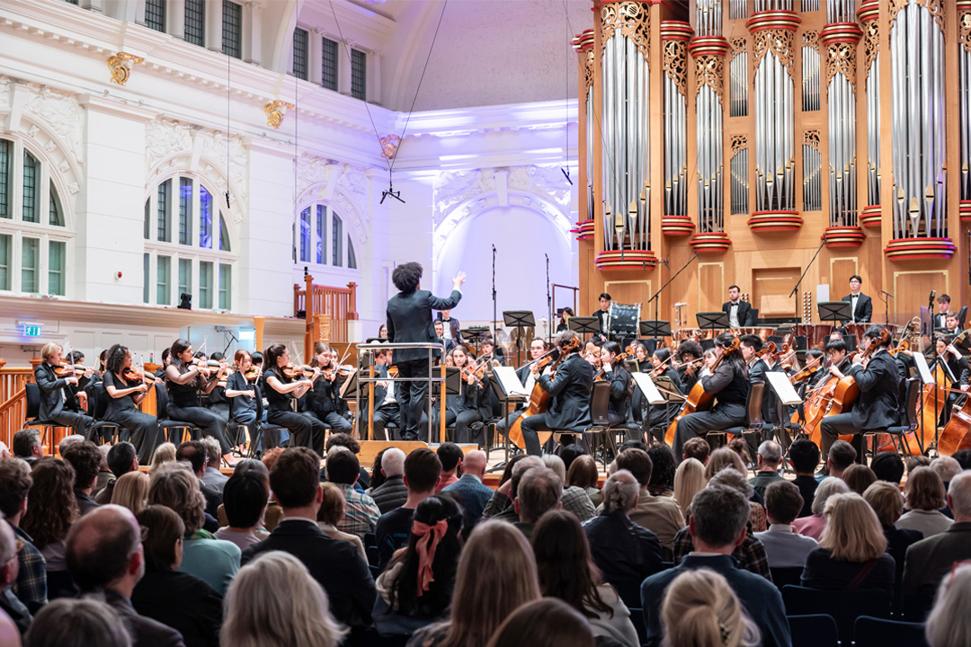 An audience watching a conductor leading an orchestra performance on stage with an organ in the background.